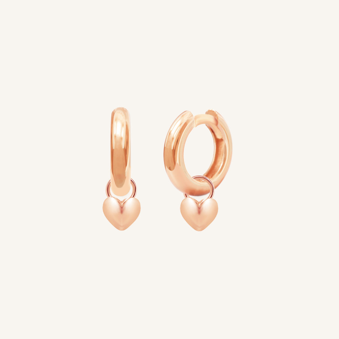 A pair of rose-colored hoop earrings with a dangling heart charm.