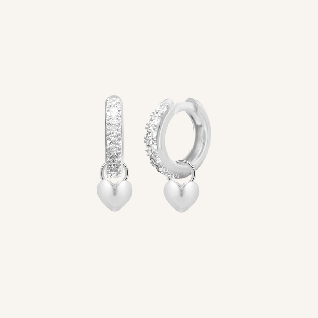 A pair of silver hoop earrings with cubic zirconias with heart charms attached to them, displayed against a white background.
