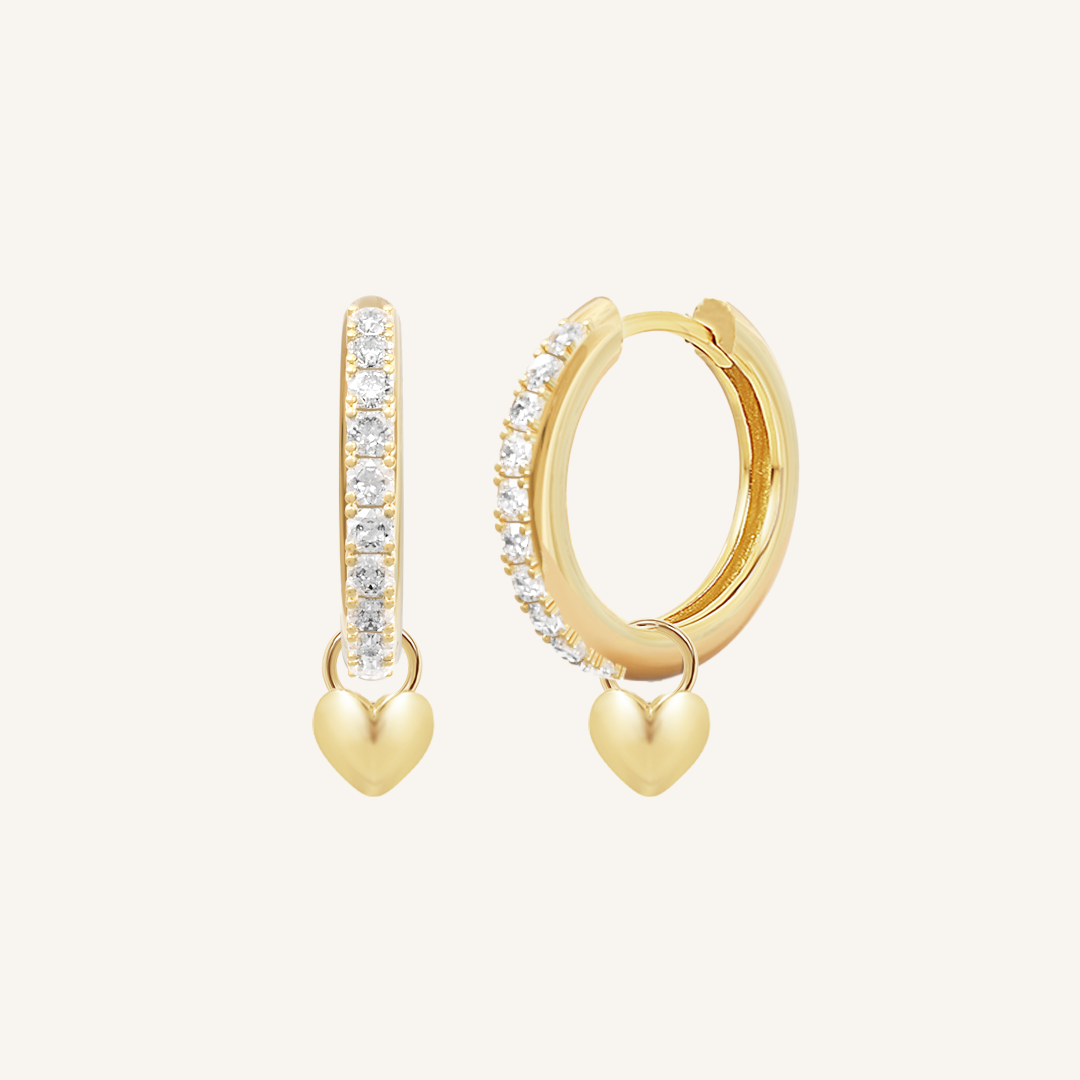 A pair of gold hoop earrings with cubic zirconias with heart charms attached to them, displayed against a white background.