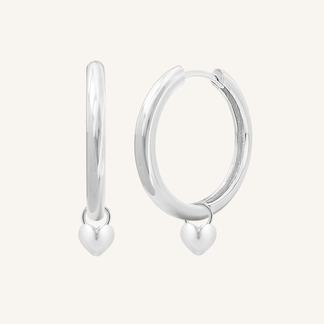 A pair of silver-colored hoop earrings with a dangling heart charm.