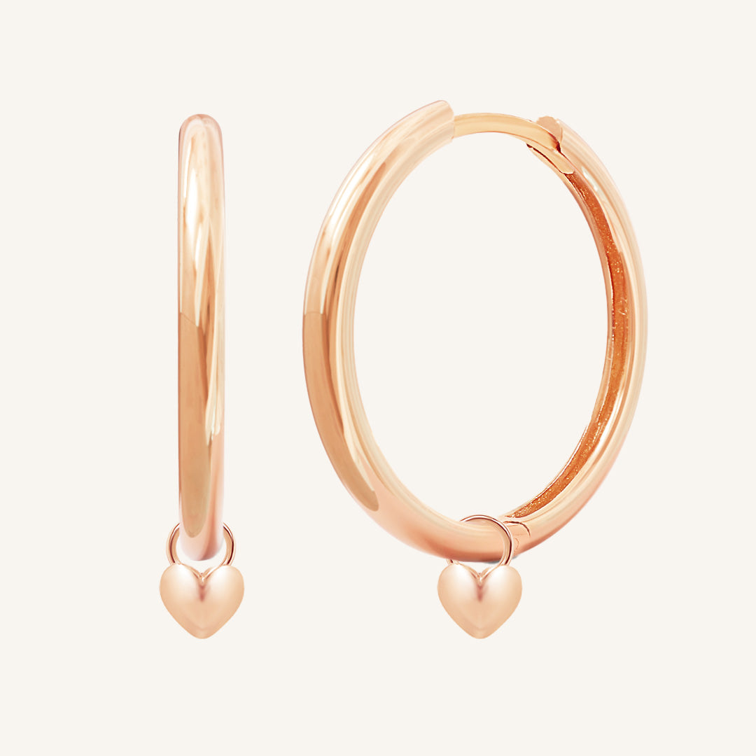 A pair of rose-colored hoop earrings with a dangling heart charm.