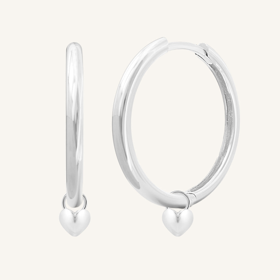 A pair of silver-colored hoop earrings with a dangling heart charm.
