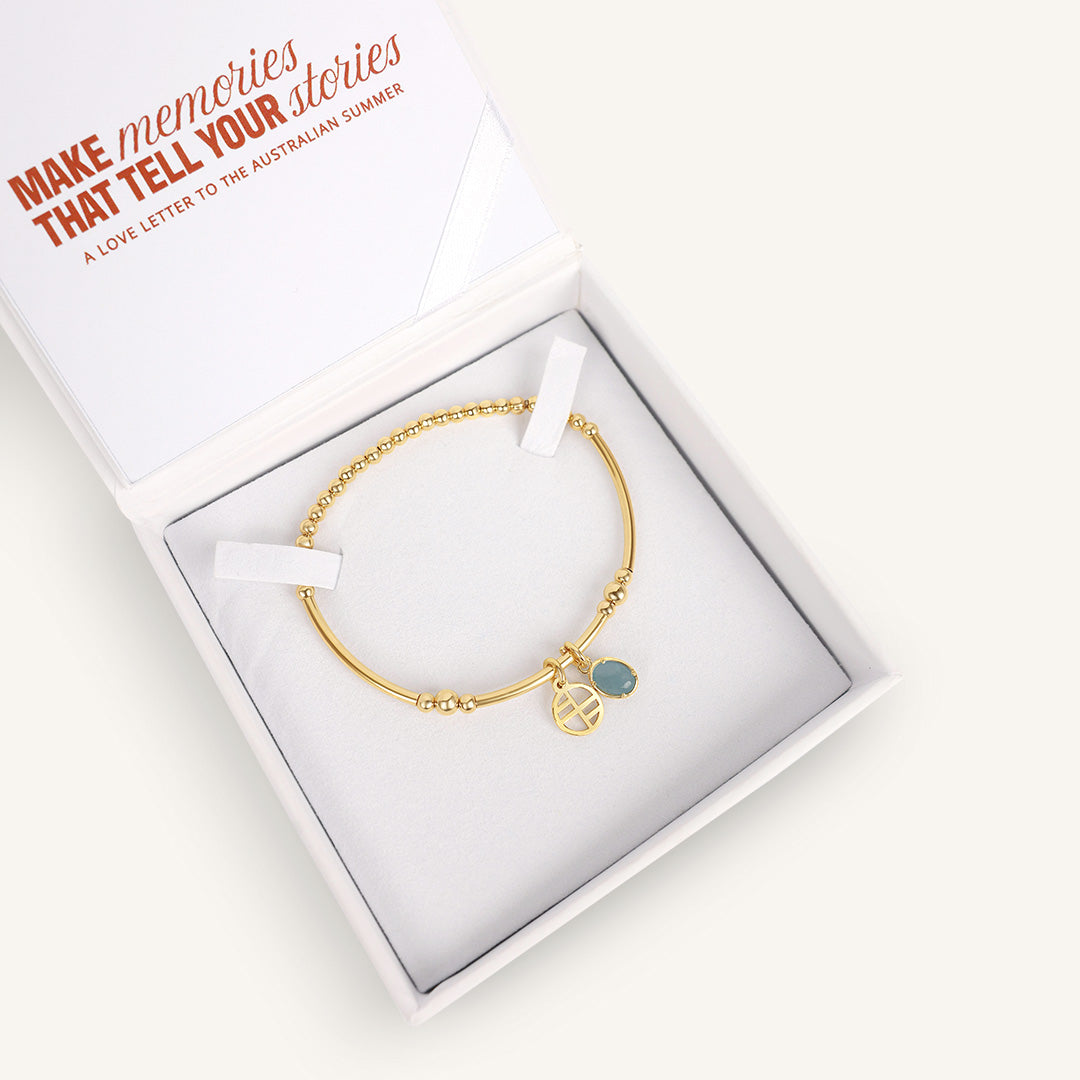 Gold bracelet with a blue charm in a white box with text on a white background