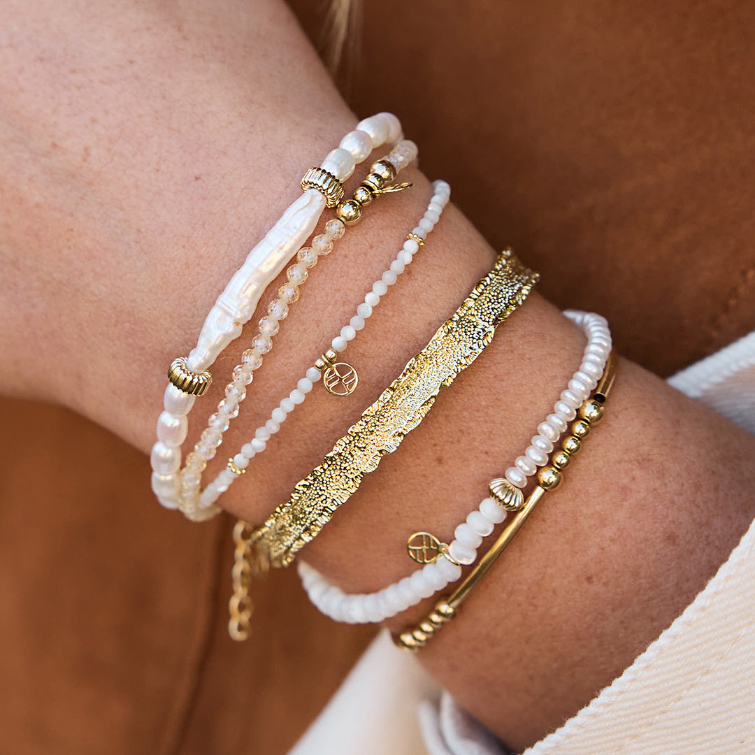 Close-up of a wrist wearing multiple gold and white beaded bracelets against a blurred background.