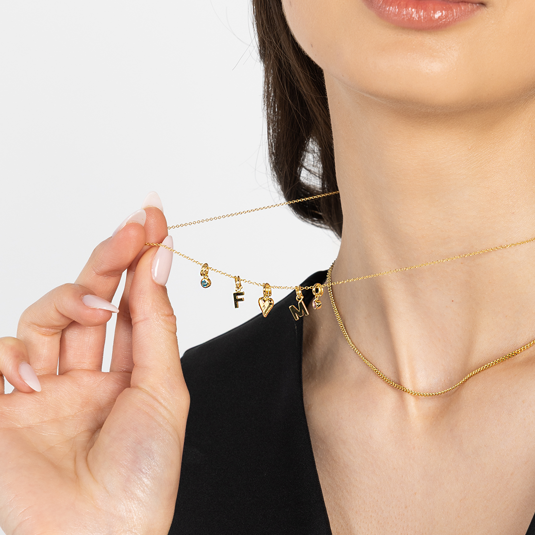 A gold-tone heart charm necklace with a fine chain with other charms, on neck, held by model