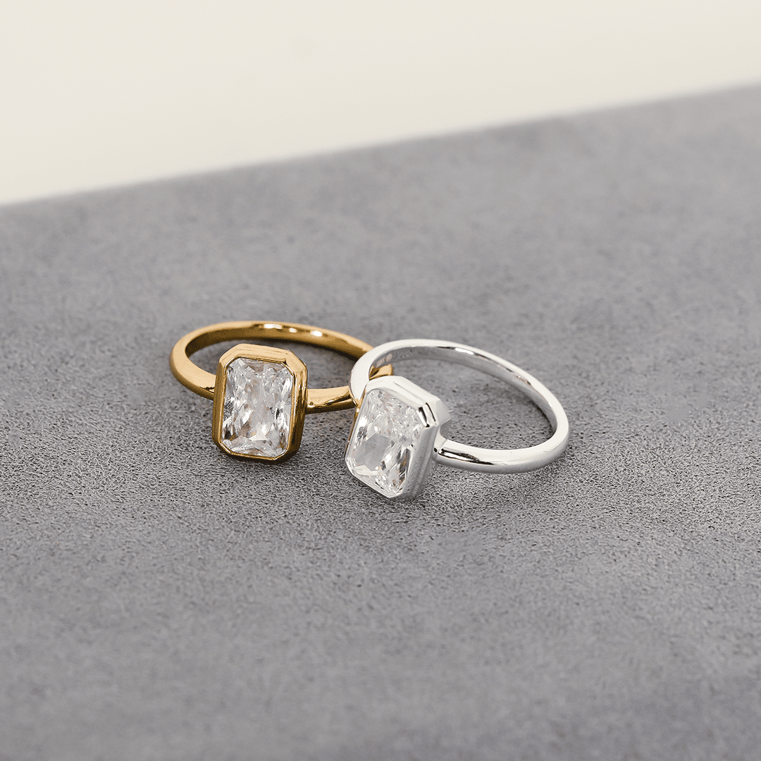 A gold-coloured and silver-coloured ring with a white cubic zirconia, bezel set in a rectangular frame with a smooth, polished band.
