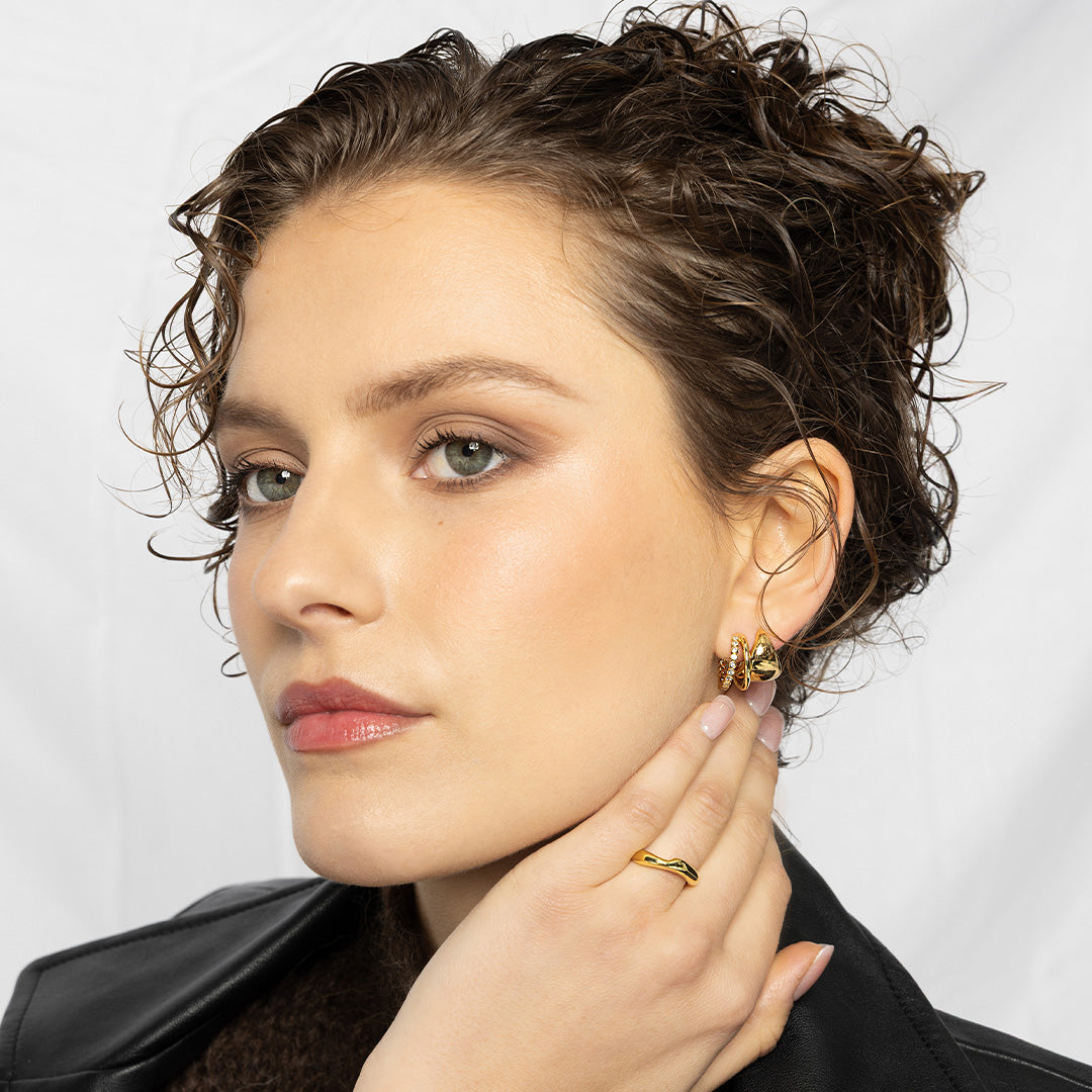 Woman wearing gold earrings and ring, touching her ear against a white background