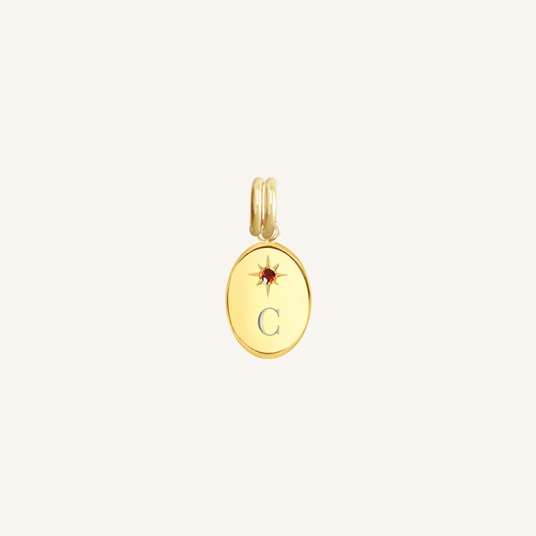 Gold pendant with letter 'C' and red stone on a light beige background