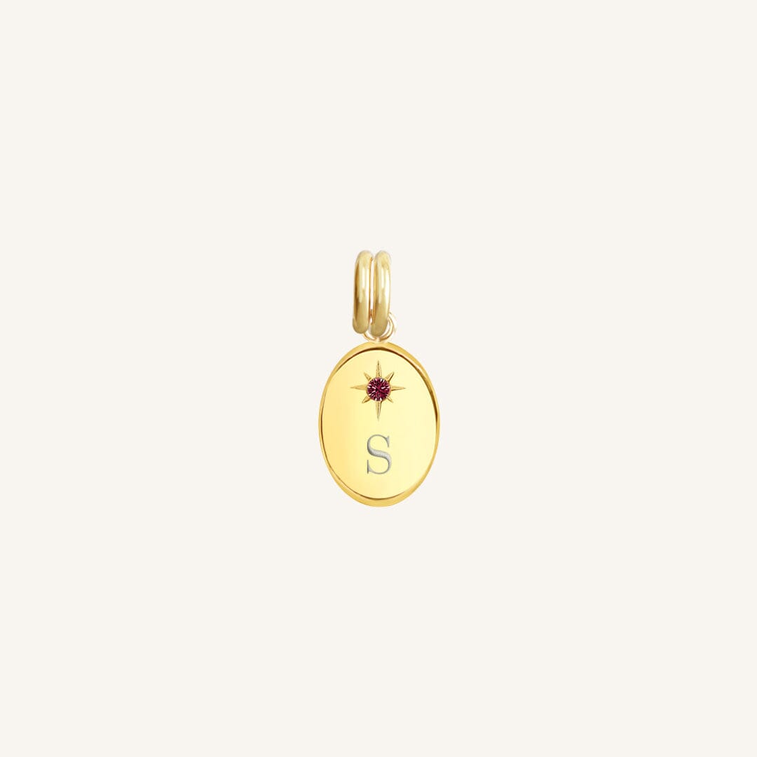 Gold pendant with a letter 'S' and red gemstone on a light beige background