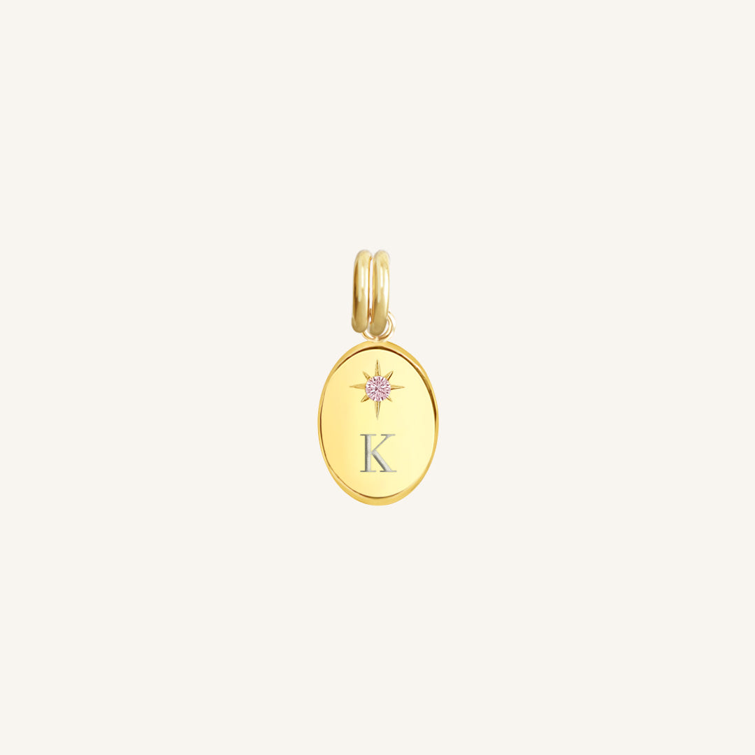 Gold pendant with 'K' and pink gemstone on a light beige background