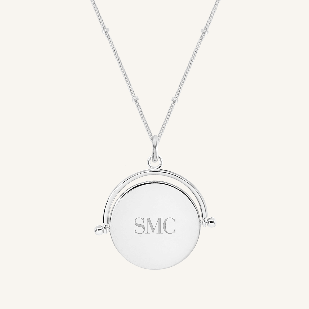 A silver-coloured pendant with the initials engraved on it, featuring a spinning mechanism, on gold chain
