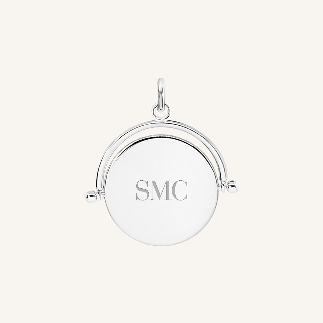 A silver-coloured pendant with the initials engraved on it, featuring a spinning mechanism.