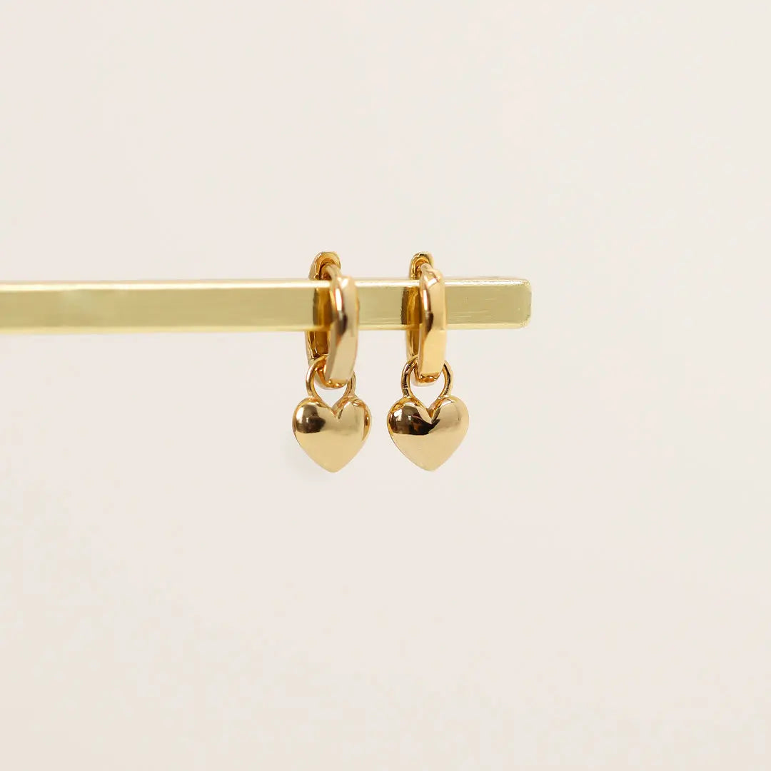 A pair of gold-colored hoop earrings with a dangling heart charm.