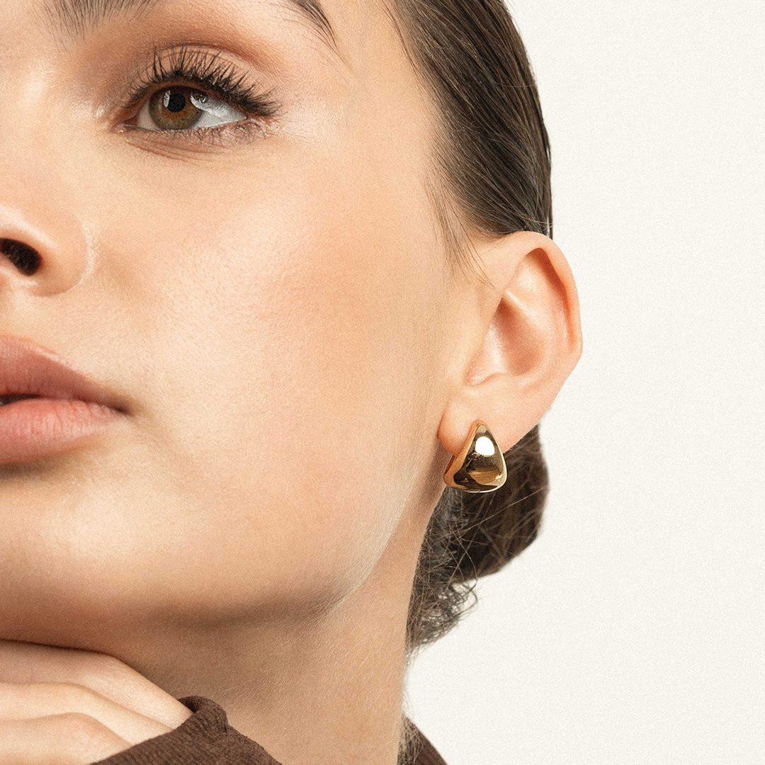 A pair of gold-colored huggie hoop earrings, worn in models ears