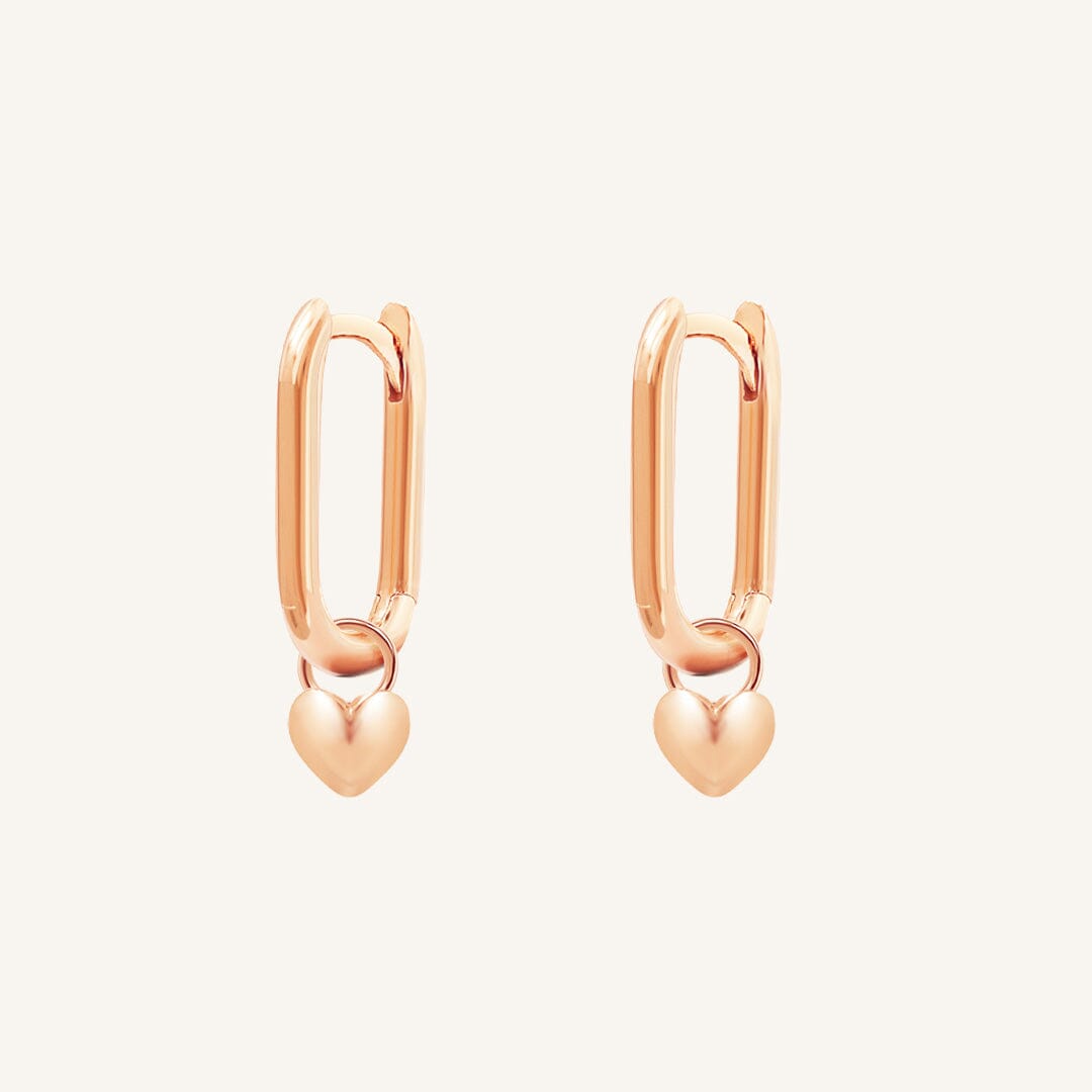 A pair of rose-tone hoop earrings with heart charms attached to them, displayed against a white background.
