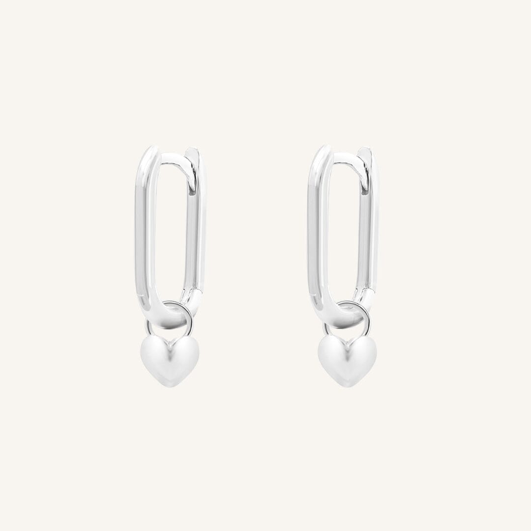 A pair of silver-tone hoop earrings with heart charms attached to them, displayed against a white background.