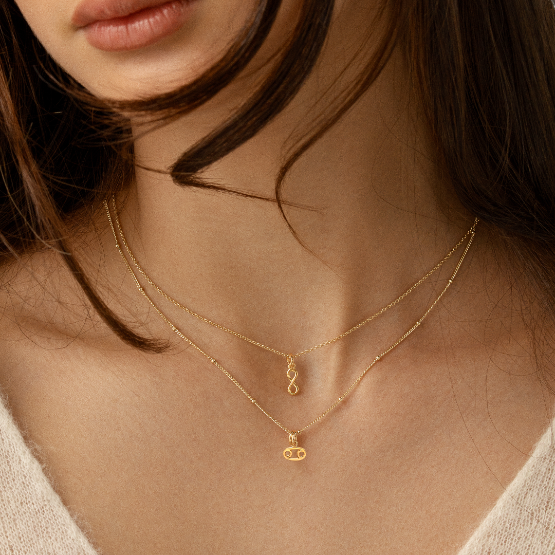 A gold-coloured Petite Zodiac Charm representing the Cancer sign on a chain around the neck