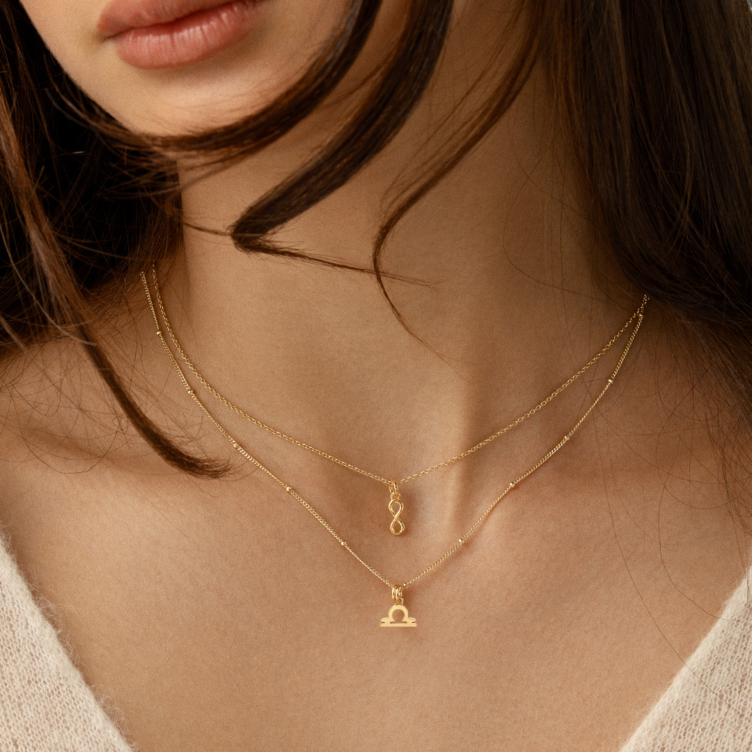 A gold-coloured Petite Zodiac Charm representing the Libra sign on a chain around the neck
