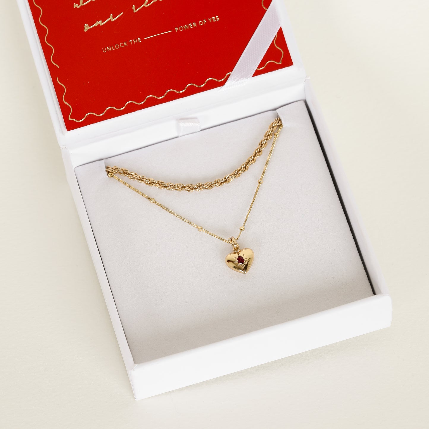 gold heart shaped charm with deep red oval cubic zirconia on bobble chain next to rope chain in white box