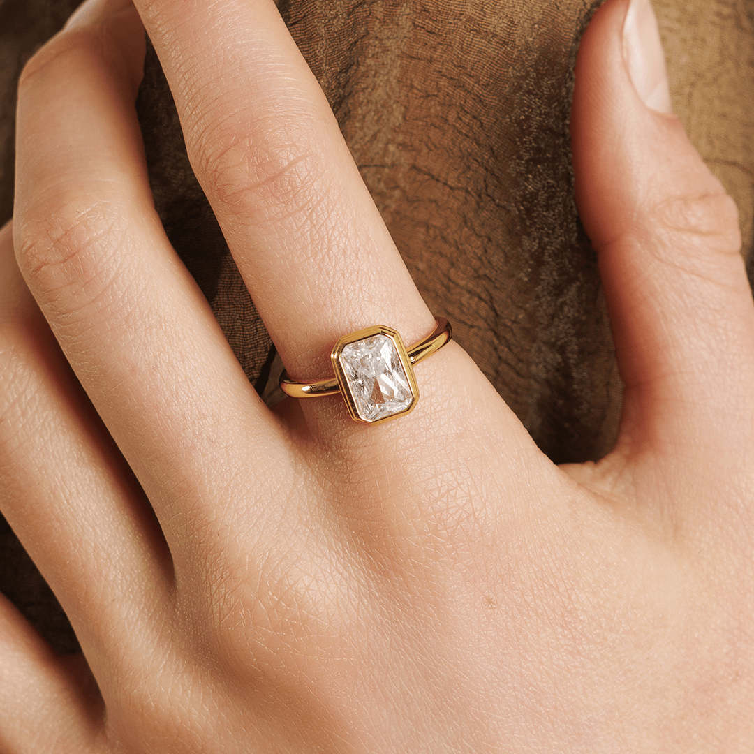 A gold-coloured ring with a white cubic zirconia, bezel set in a rectangular frame with a smooth, polished band worn on finger