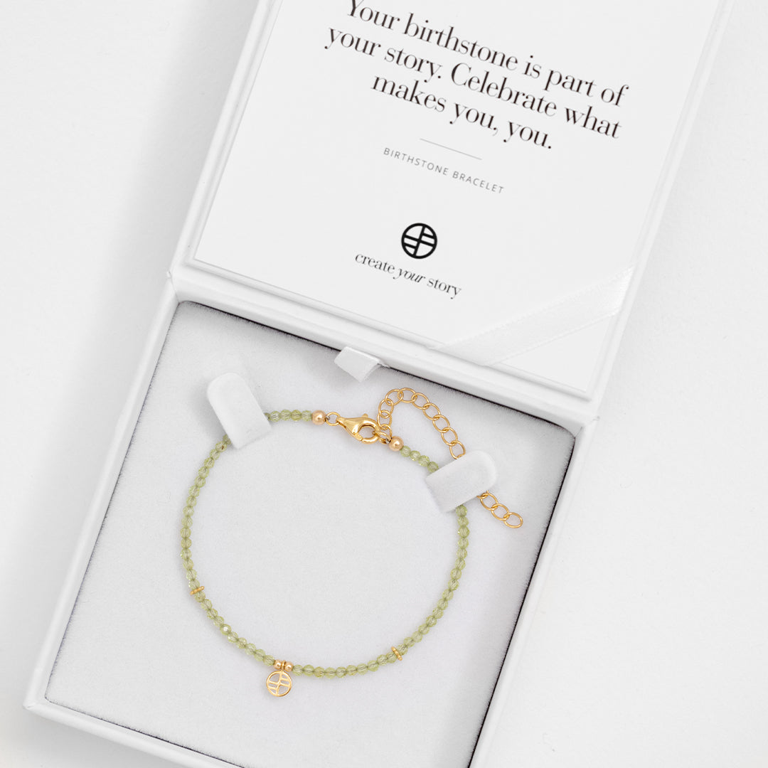 A beaded bracelet with green stones and a gold metal clasp, representing the August birthstone, in white box