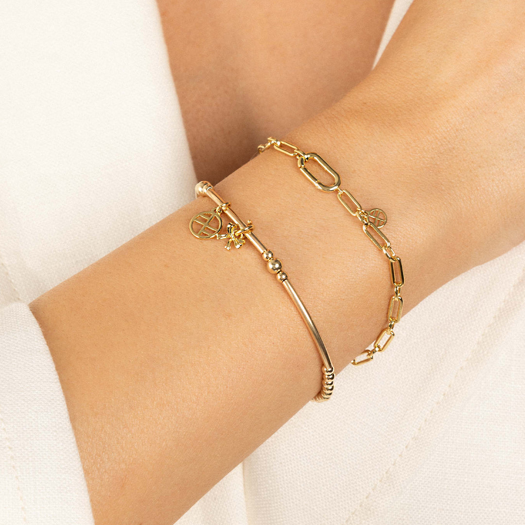 A gold-tone metal bracelet with a beaded design, featuring a charm in the shape of a bow, worn on wrist