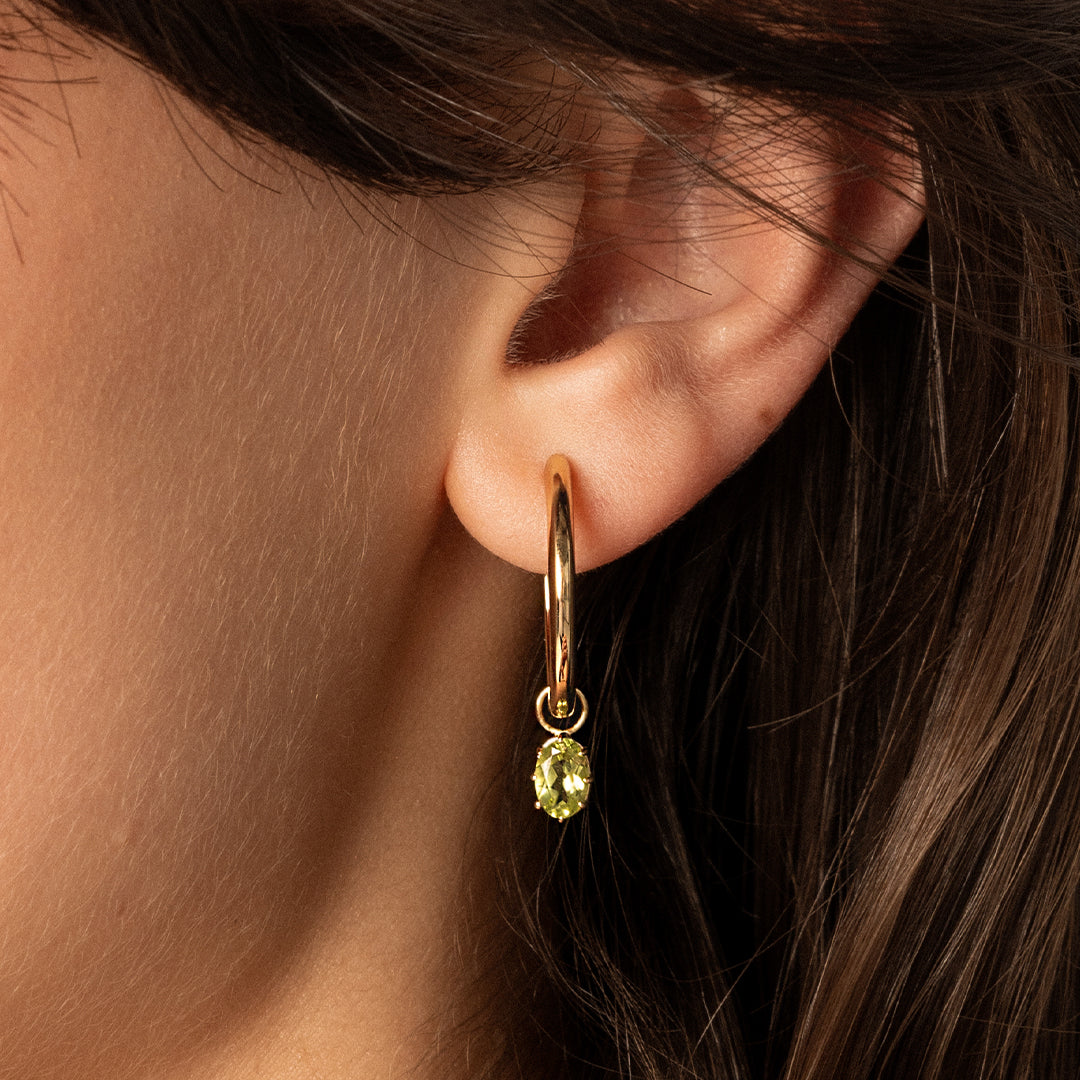 A pair of gold-tone hoop earrings with olive green charms attached to them, close up in ears