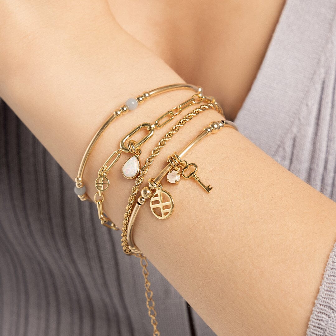 A gold-tone charm bracelet featuring a beaded design with a small circular charm and a moonstone charm, worn on models wrist, layered with other bracelets