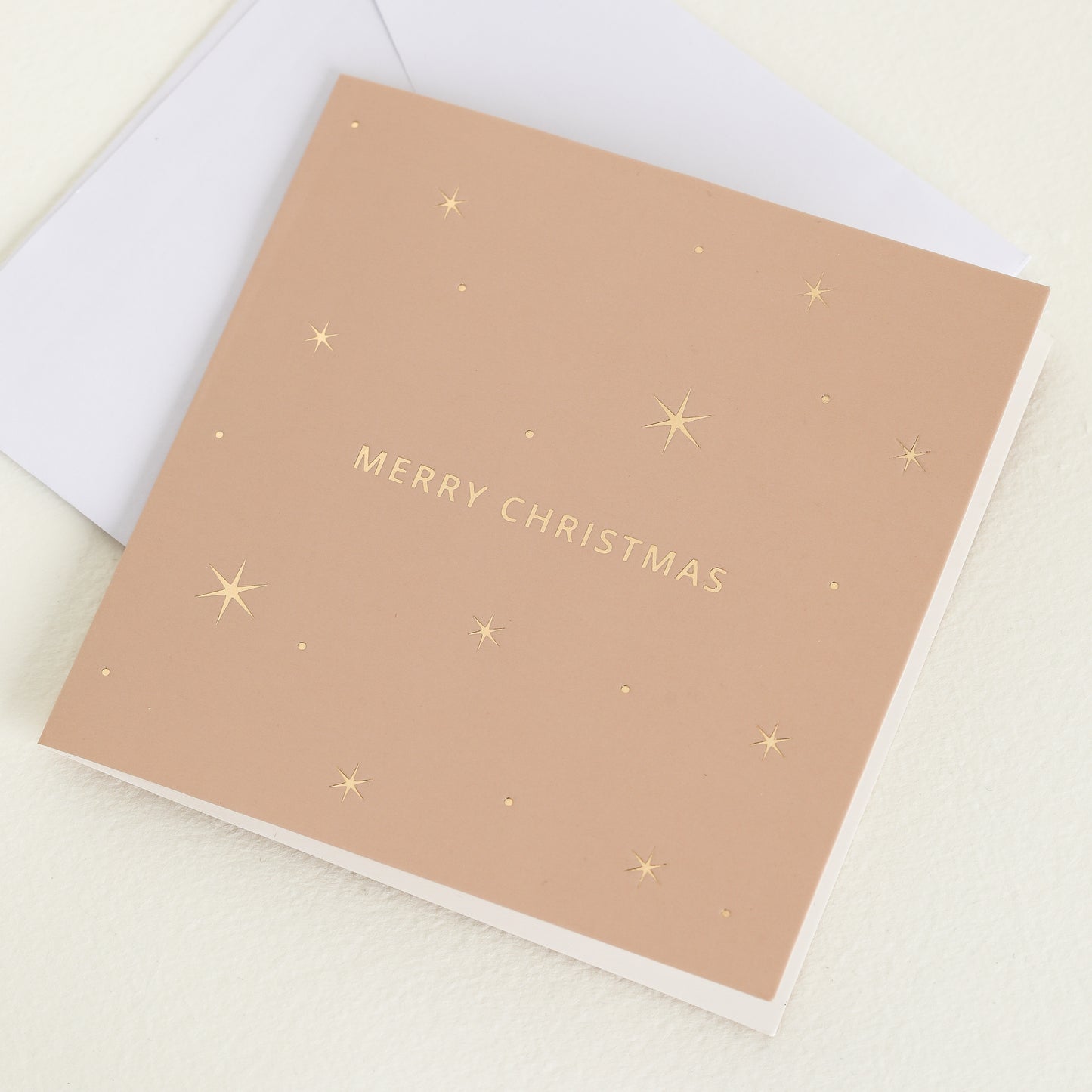 A pink greeting card with the word 'MERRY CHRISTMAS' printed in gold letters, accompanied by a white envelope.