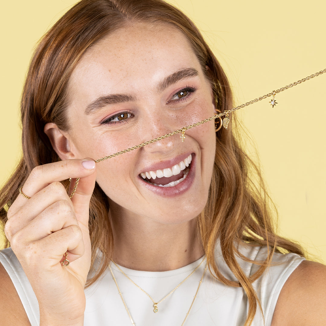 A petite gold cross charm with cubic zirconia on gold chain, being held by model