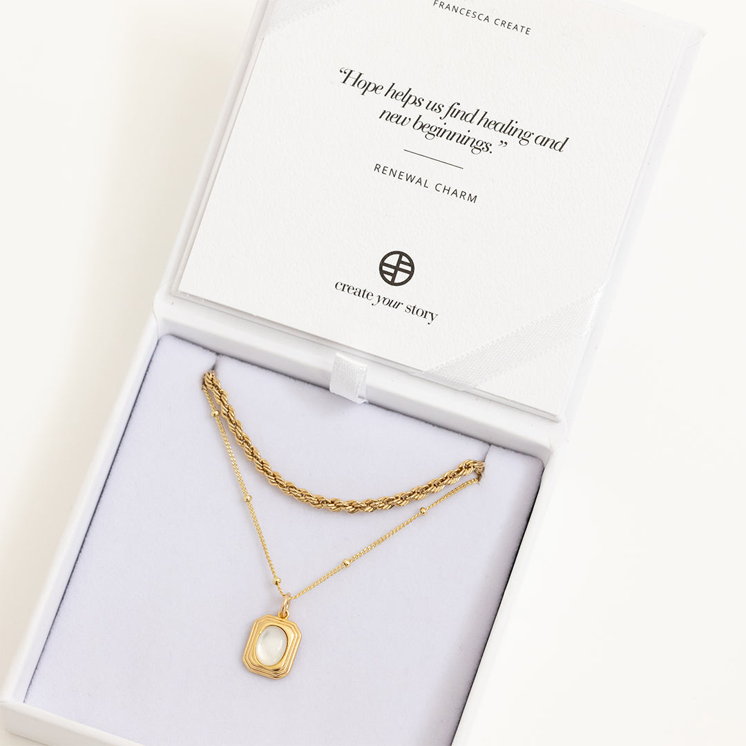 A gold-colored pendant with an oval mother-of-pearl stone in the center, on bobble chain, displayed in white box