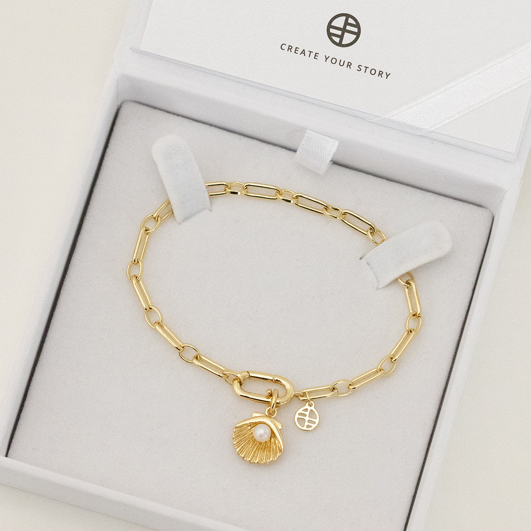 A gold-colored chain bracelet with a shell shaped charm with a pearl in the middle, displayed in white box