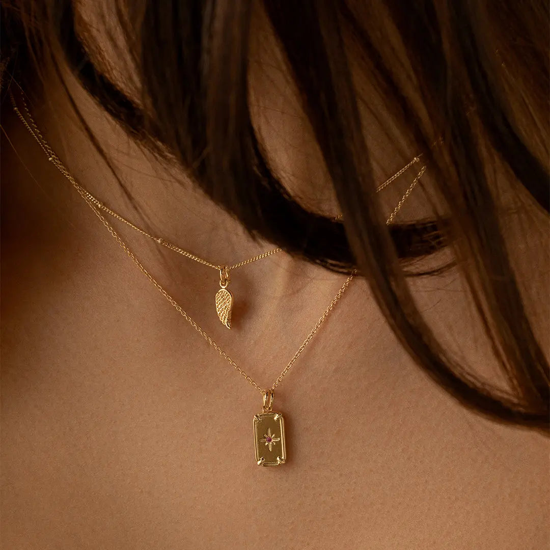 A gold-coloured angel wing charm with intricate details, on a necklace, on models neck