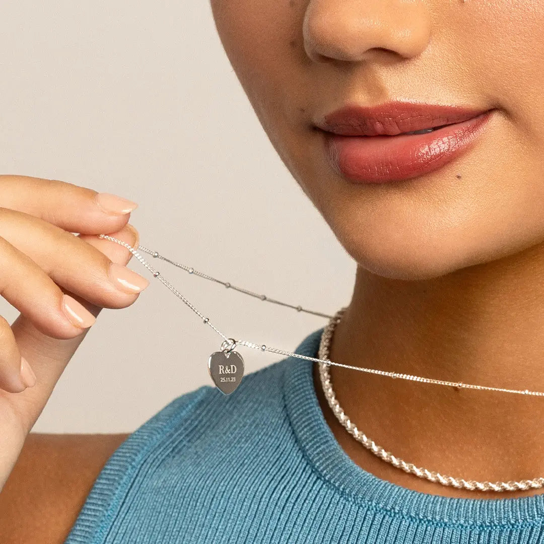 A silver heart-shaped charm with engraved initials and a date, worn on silver chain, worn on models neck