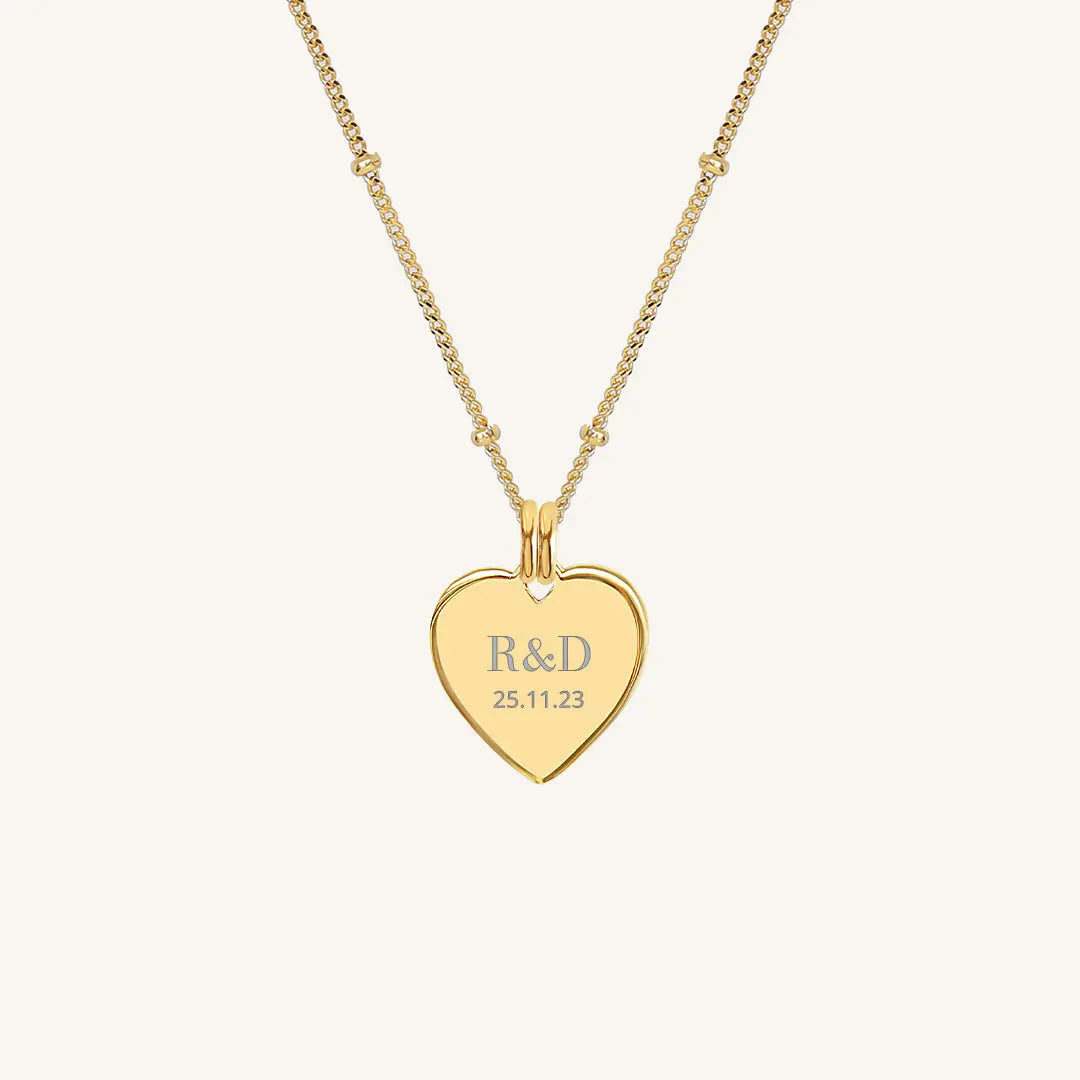 A gold heart-shaped necklace with a chain, featuring an engraved monogram initials and a date on the front.