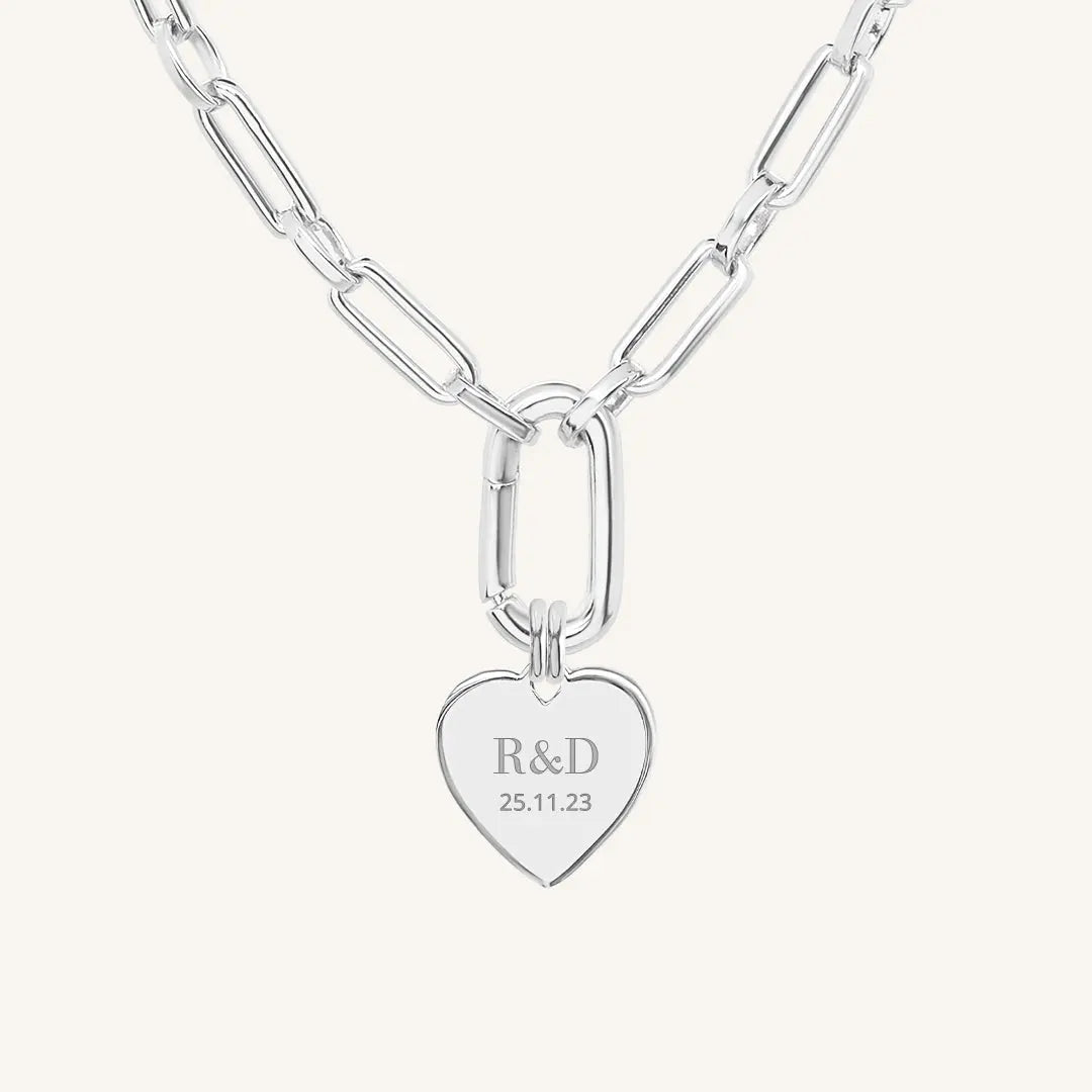 A silver heart-shaped necklace with a chain, featuring an engraved monogram initials and a date on the front.