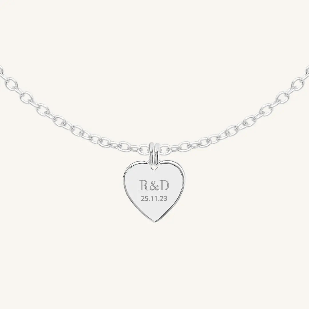 A silver heart-shaped necklace with a chain, featuring an engraved monogram initials and a date on the front.