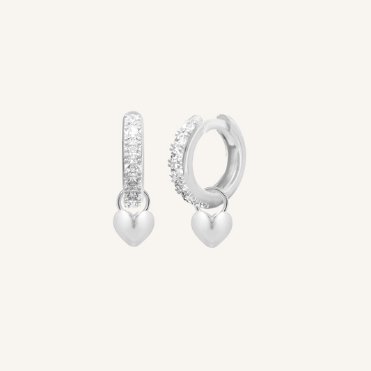A pair of silver hoop earrings with cubic zirconias with heart charms attached to them, displayed against a white background.