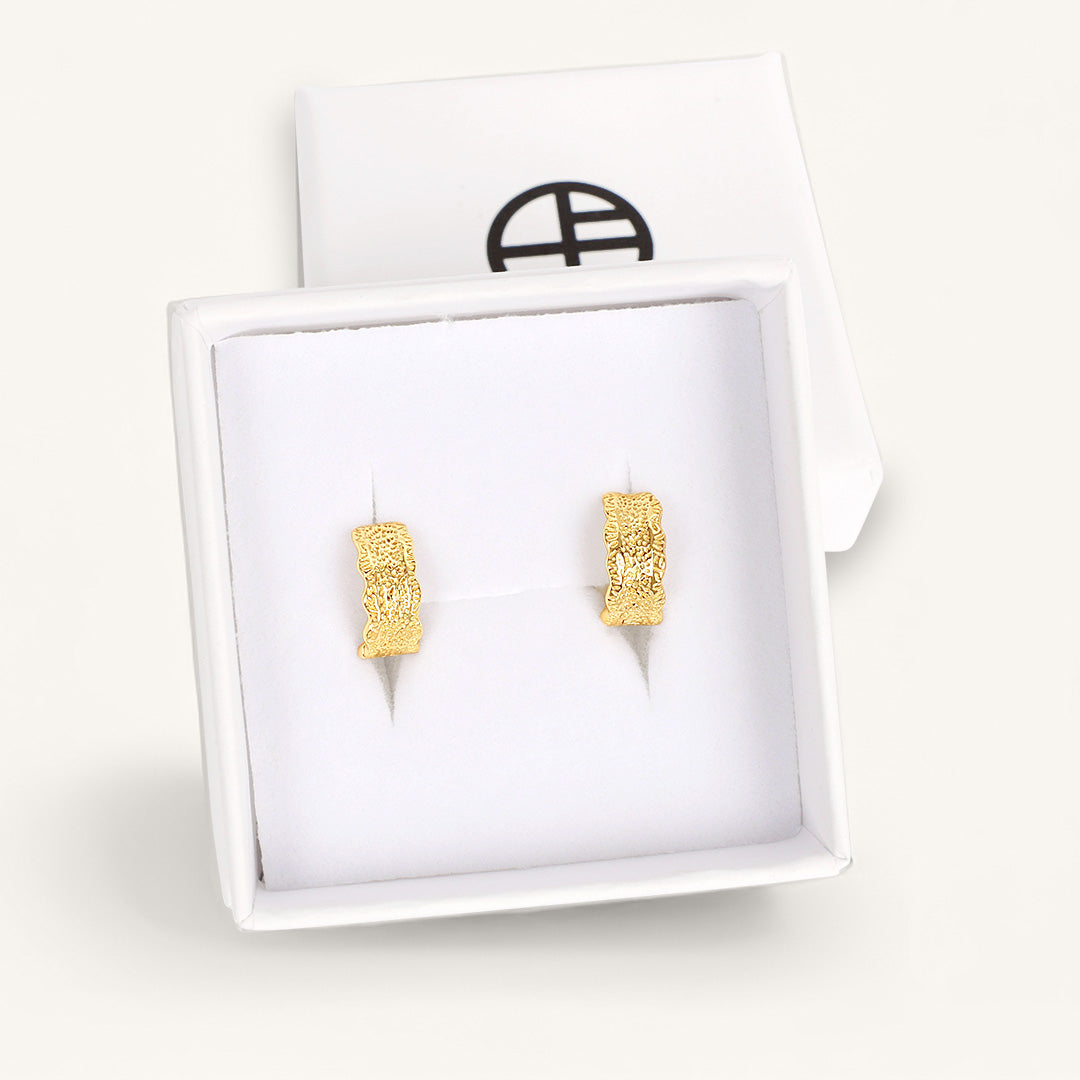 Gold textured square earrings in a white display box with a brand logo in the background.