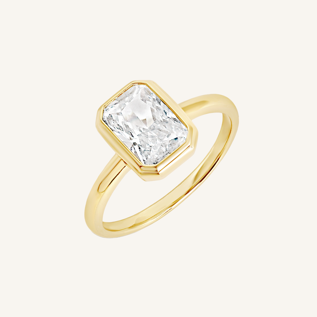 A gold-coloured ring with a white cubic zirconia, bezel set in a rectangular frame with a smooth, polished band.