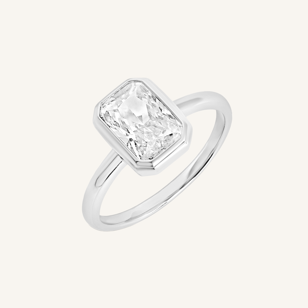 A silver-coloured ring with a white cubic zirconia, bezel set in a rectangular frame with a smooth, polished band.