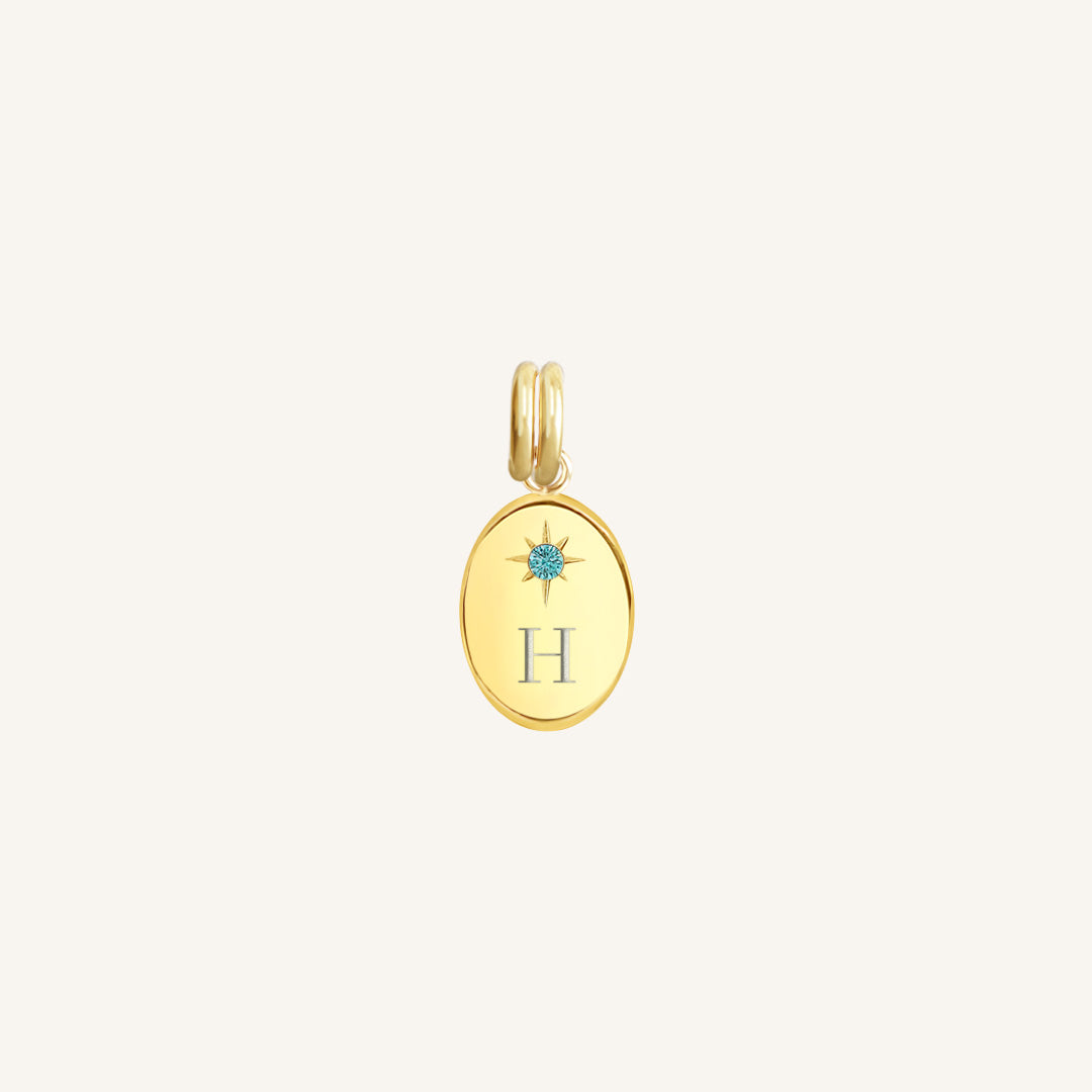 Gold pendant with 'H' and blue gemstone on a light beige background