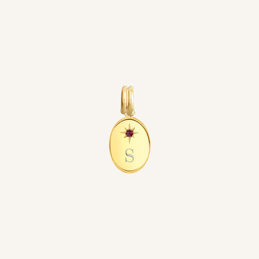 Gold pendant with a letter 'S' and red gemstone on a light beige background