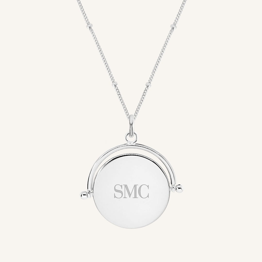 A silver-coloured pendant with the initials  engraved on it, featuring a spinning mechanism, on gold chain