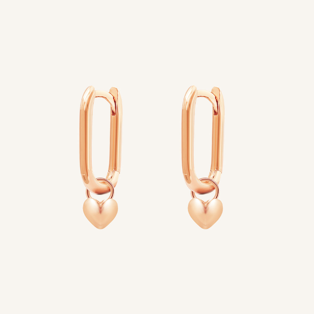 A pair of rose-tone hoop earrings with heart charms attached to them, displayed against a white background.