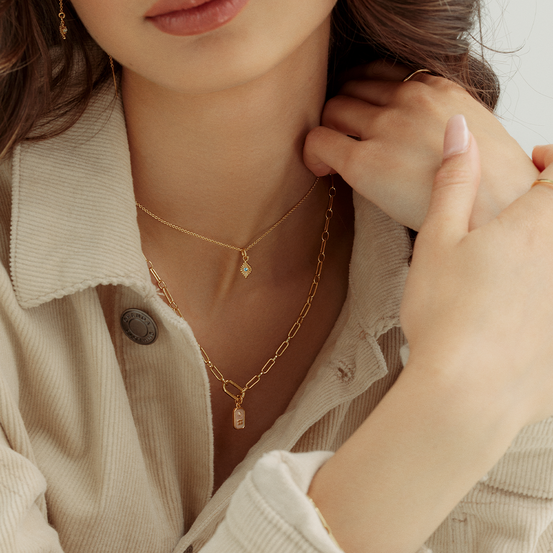 A gold charm with a green cubic zirconia, resembling a May birthstone, worn on gold chain, on models neck.