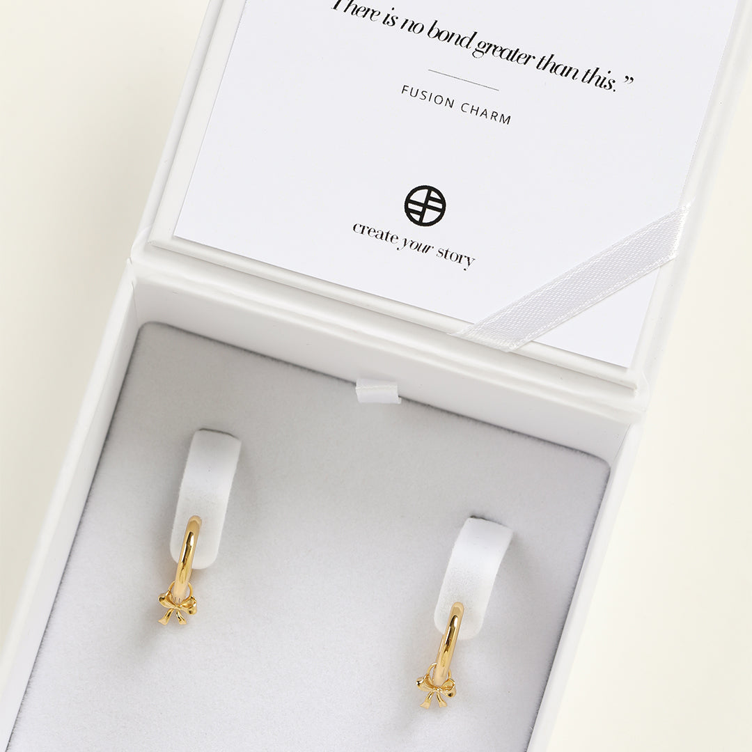 A set of two gold hoop charms with a bow design, on hoops, in white box
