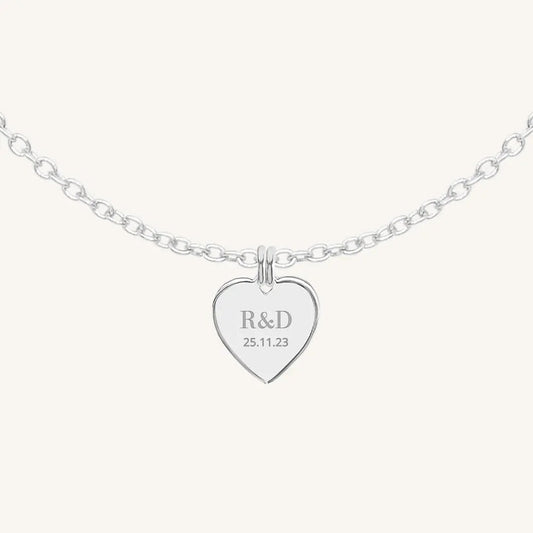 A silver heart-shaped necklace with a chain, featuring an engraved monogram initials and a date on the front.
